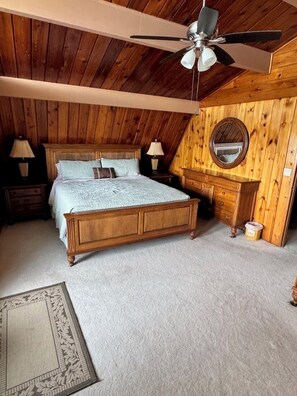 A bedroom with a wooden ceiling, a bed, a nightstand, a dresser, a mirror, and a ceiling fan. - The Mossy Gambrel on Lake Huntley- fish, ski, swim, kayak and relax by fire pit! (Lake Placid)