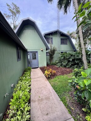 Un edificio verde con una puerta gris, un pequeño jardín y un cielo despejado.