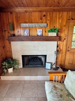 A wooden cabin interior with a fireplace, a sign above it, and a door to the right. - The Mossy Gambrel on Lake Huntley- fish, ski, swim, kayak and relax by fire pit! (Lake Placid)