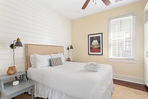 A neatly arranged bedroom with a bed, bedside tables, a wall-mounted lamp, a framed picture, and a window with blinds. - Mint Julep Residence @ Smalls Alley w/ Heated Saltwater Pool (Charleston)