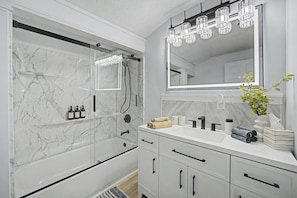 Un baño moderno con ducha envuelta en vidrio, paredes de mármol, un espejo grande y un tocador blanco con lavamanos y perchero.