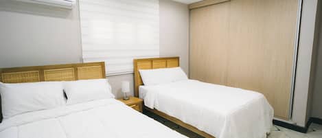 A hotel room with two single beds, a bedside table with a lamp, a window with blinds, and an air conditioning unit on the wall.