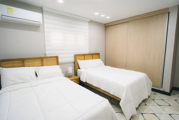 A hotel room with two single beds, a bedside table with a lamp, a window with blinds, and an air conditioning unit on the wall.