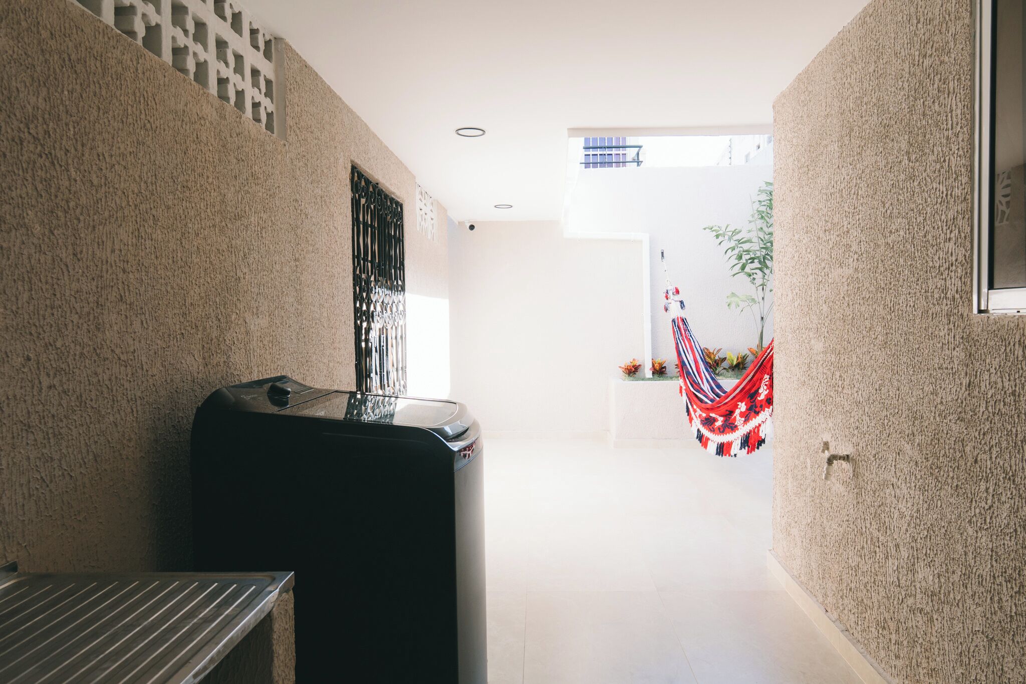 A narrow hallway with a trash bin, a window with a metal grille, and a hammock hanging in the distance.