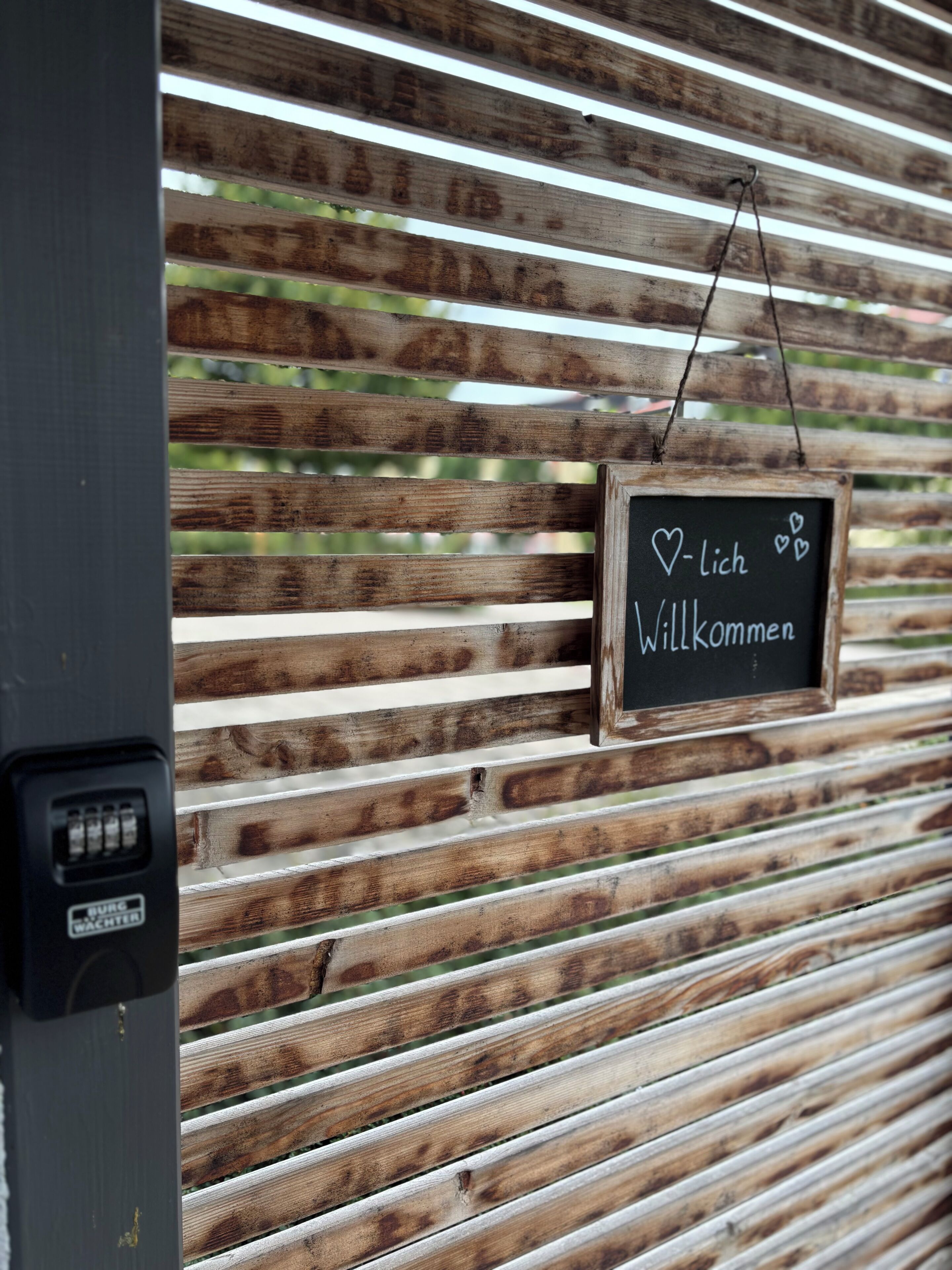 Un mur en bois à lattes sur lequel est fixé un tableau noir où l’on peut lire le mot « WILLKOMMEN » et des symboles en forme de cœur.