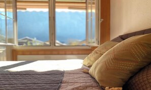 A bed with a quilt and two pillows, a window with a view of mountains, and a wooden headboard. - BEAUTIFUL 2-BEDROOM CHALET WITH PANORAMIC MOUNTAIN VIEWS (Lauterbrunnen)