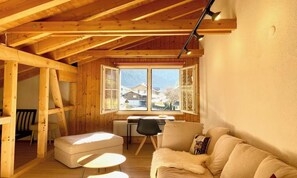 A cozy room with a wooden ceiling, a sofa, a small table, and a window with a view of houses. - BEAUTIFUL 2-BEDROOM CHALET WITH PANORAMIC MOUNTAIN VIEWS (Lauterbrunnen)