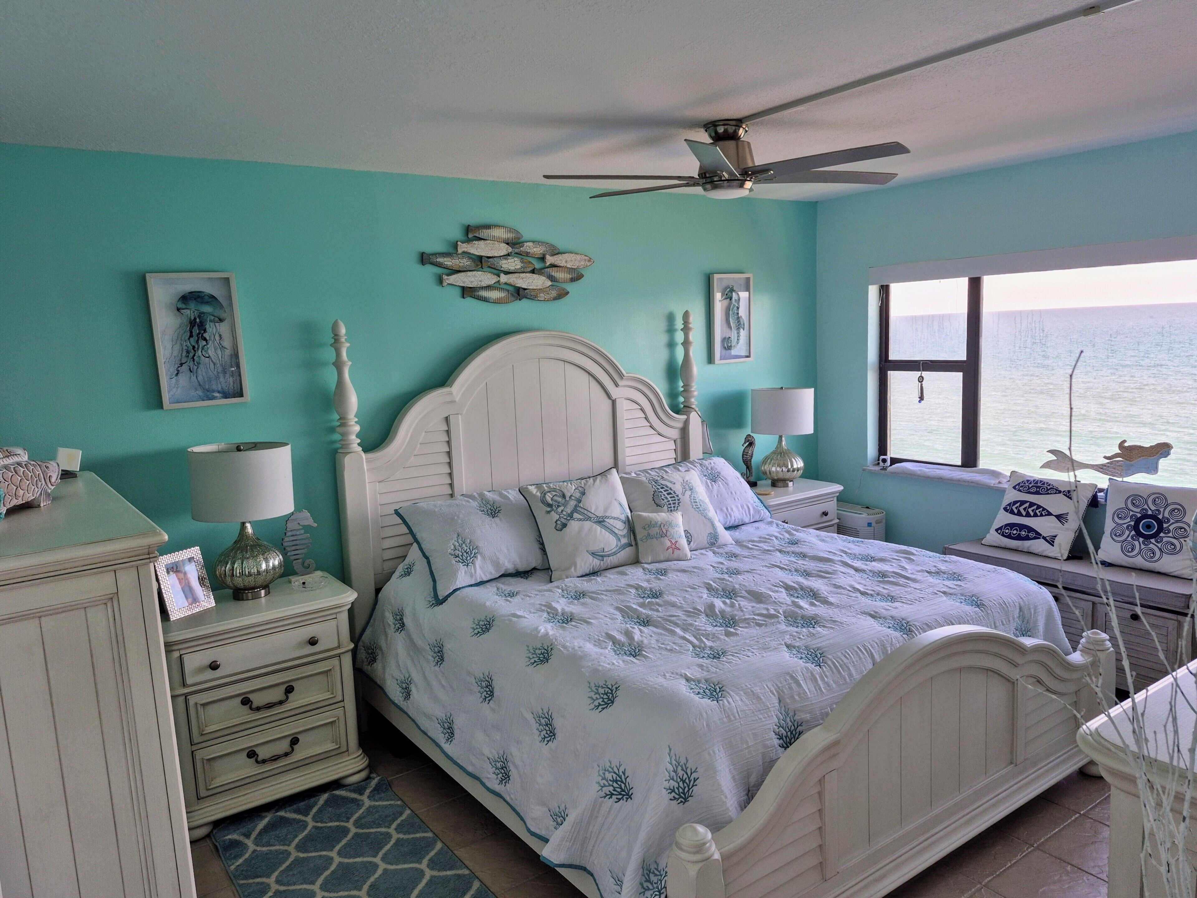 Een slaapkamer met een bed, nachtkastjes, een dressoir, een plafondventilator en een raam met uitzicht op zee.