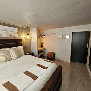 A modern bedroom with a large bed, bedside tables, a hanging light fixture, a small bench, and a dark door. - Modern hidden retreat with chef’s kitchen, spa bath, pergola, bar & solar-power. (Boquerón)