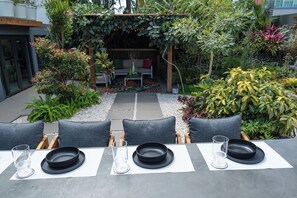 An outdoor dining area with a table set for four, surrounded by chairs, with a garden and a building in the background. - Modern hidden retreat with chef’s kitchen, spa bath, pergola, bar & solar-power. (Boquerón)