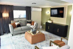 Een moderne woonkamer met een grijze bank, een bruin poefje, een zwart kastje en een flatscreen aan de muur.