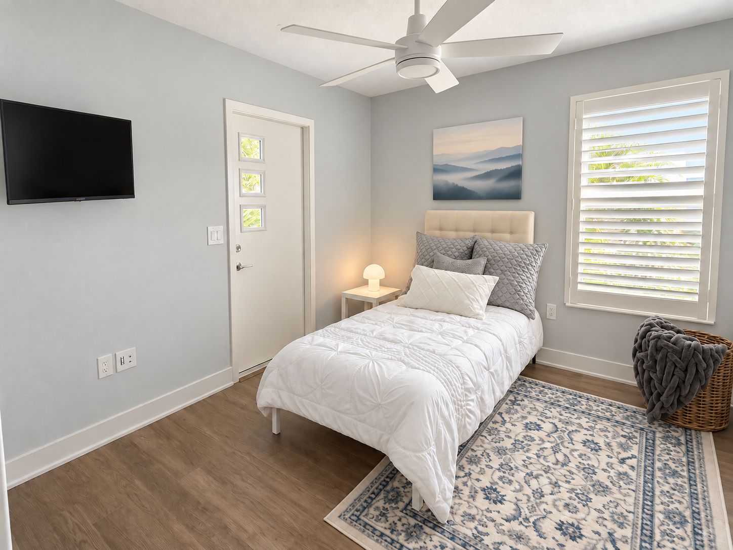 A bedroom with a bed, a ceiling fan, a wall-mounted TV, a door with a window, and a window with blinds.