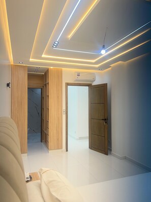A modern hotel room with a clean, minimalist design, featuring a sofa, a wardrobe, and a ceiling with recessed lighting.