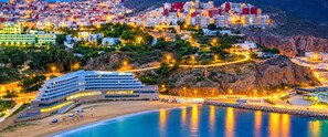 A coastal hotel with multiple floors, a beach, and a rocky hillside in the background.