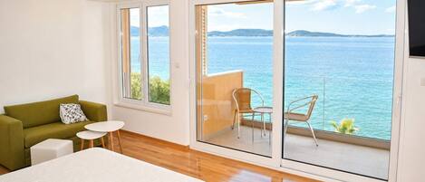 A room with a bed, a green sofa, a small table, and a balcony with a view of the sea.