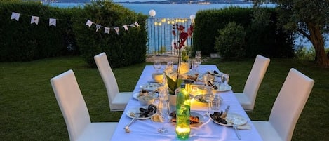 Un montaje para comer al aire libre con una mesa lista para una comida, rodeada de sillas, y una vista al mar y colinas en el fondo.