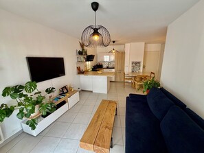 Ein modernes Wohnzimmer mit Fernseher, Couchtisch aus Holz, blauem Sofa und einer Küchenzeile mit Holzschränken und einem Esstisch.