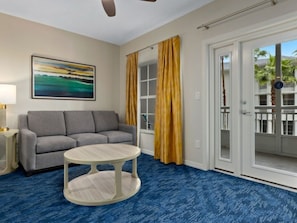 Ein Hotelzimmer mit grauem Sofa, einem runden Tisch, einem Gemälde an der Wand und Blick auf ein Gebäude mit Balkonen.