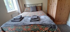 Un llit amb llençols florals, dues tovelloles doblegades i una finestra amb cortines.