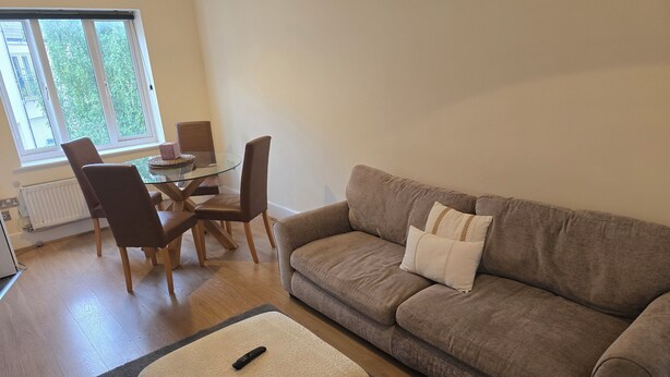 Una sala de estar con un sofá marrón, dos sillones, una mesa de centro de vidrio y una mesa de comedor con sillas.