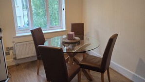Un comedor con una mesa de vidrio, cuatro sillas, un radiador y una ventana con vistas a edificios y árboles.