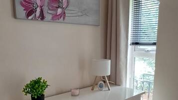 Une pièce avec une commode blanche, une petite plante, un tableau, une fenêtre avec des rideaux et un radiateur.
