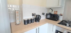 A kitchen with a coffee maker, tea kettle, and sugar and coffee containers on the counter. - Stylish yet Cosy 3-Bed Flat with Private Balcony. (Stoke Gifford)