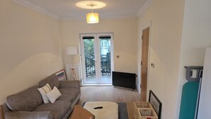Una sala de estar con sofá, reposapiés y televisor. Hay una puerta que da a un balcón.