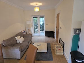 Ein Wohnzimmer mit einer Couch, einem Hocker und einem Fernseher. Eine Tür führt zu einem Balkon.