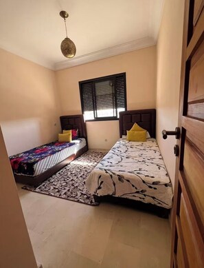 A bedroom with two beds, a window with blinds, a hanging light fixture, and a wooden door. - 2-bedroom apartment in enchanting Ouahat Sidi Brahim with AC (Ouahat Sidi Brahim)