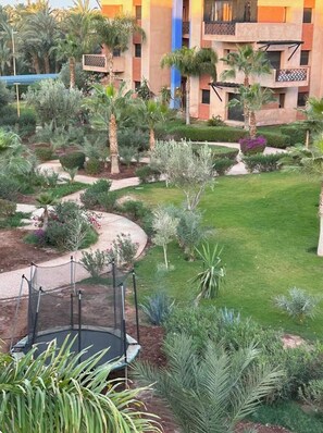 A landscaped garden with palm trees, a winding path, and a building with balconies. - 2-bedroom apartment in enchanting Ouahat Sidi Brahim with AC (Ouahat Sidi Brahim)