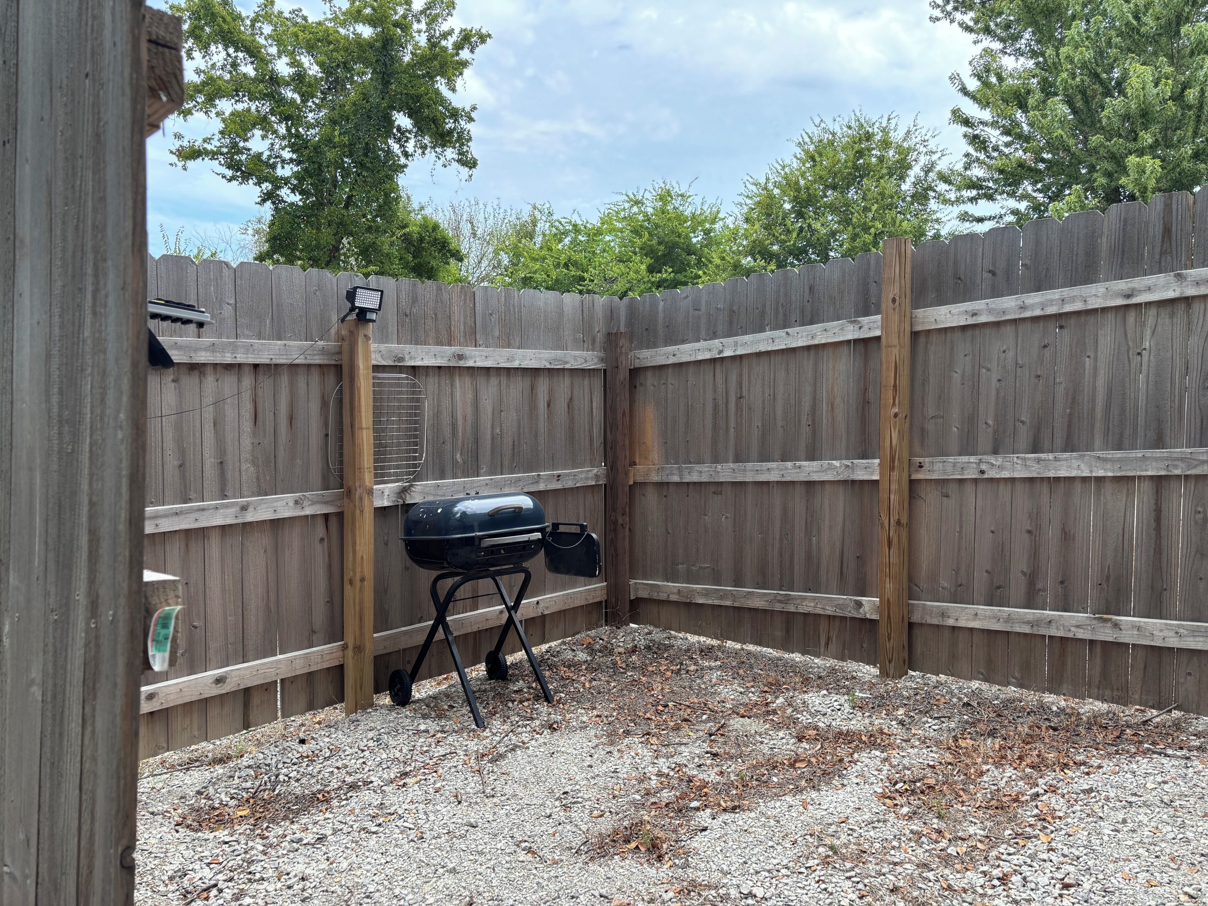 Un patio trasero con cerca de madera, una parrilla y suelo de grava.