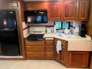 A kitchen with wooden cabinets, a black refrigerator, a microwave, a stove, a sink, and a coffee maker. - Experience a unique stay in a well appointed motorhome (Willsboro)
