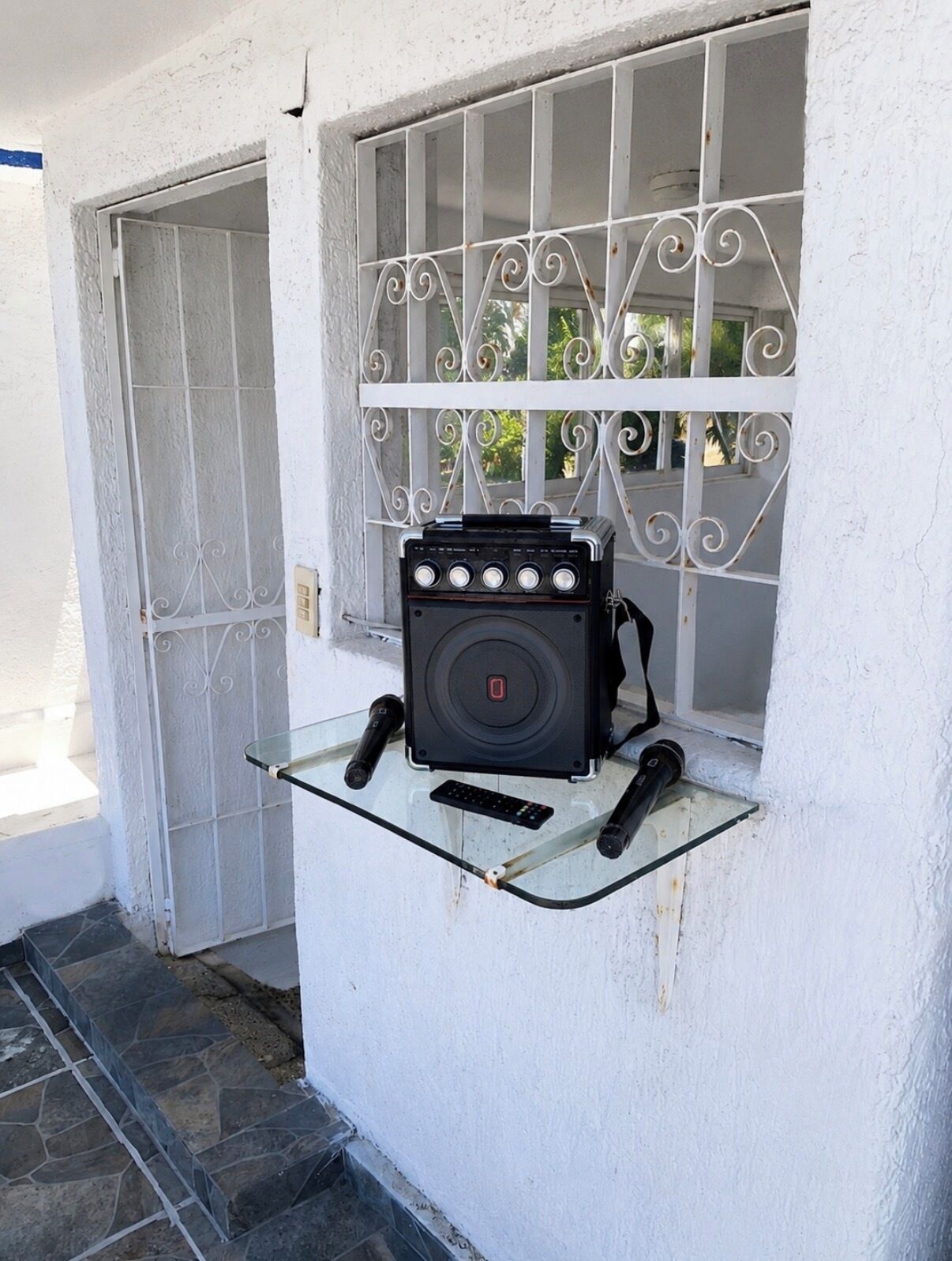 A portable gas stove on a glass shelf outside a white building with a metal gate and window.