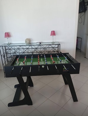 Una mesa de fútbol de mesa en una habitación con piso de baldosas y una mesa de vidrio con lámparas rosadas al fondo.
