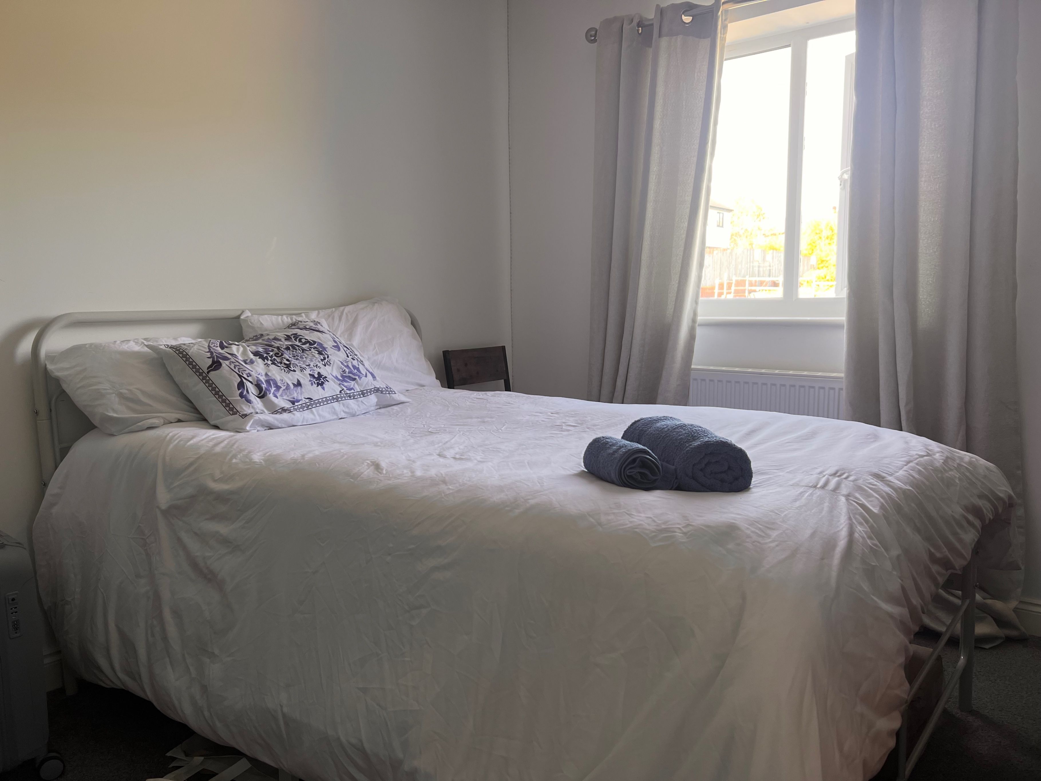 A neatly made bed with white bedding and a decorative pillow, a rolled towel on the bed, and a window with curtains.
