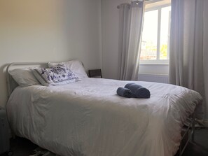 A neatly made bed with white bedding and a decorative pillow, a rolled towel on the bed, and a window with curtains. - Cozy Crook Retreat With 2 Bedrooms Near Kyren (Crook)
