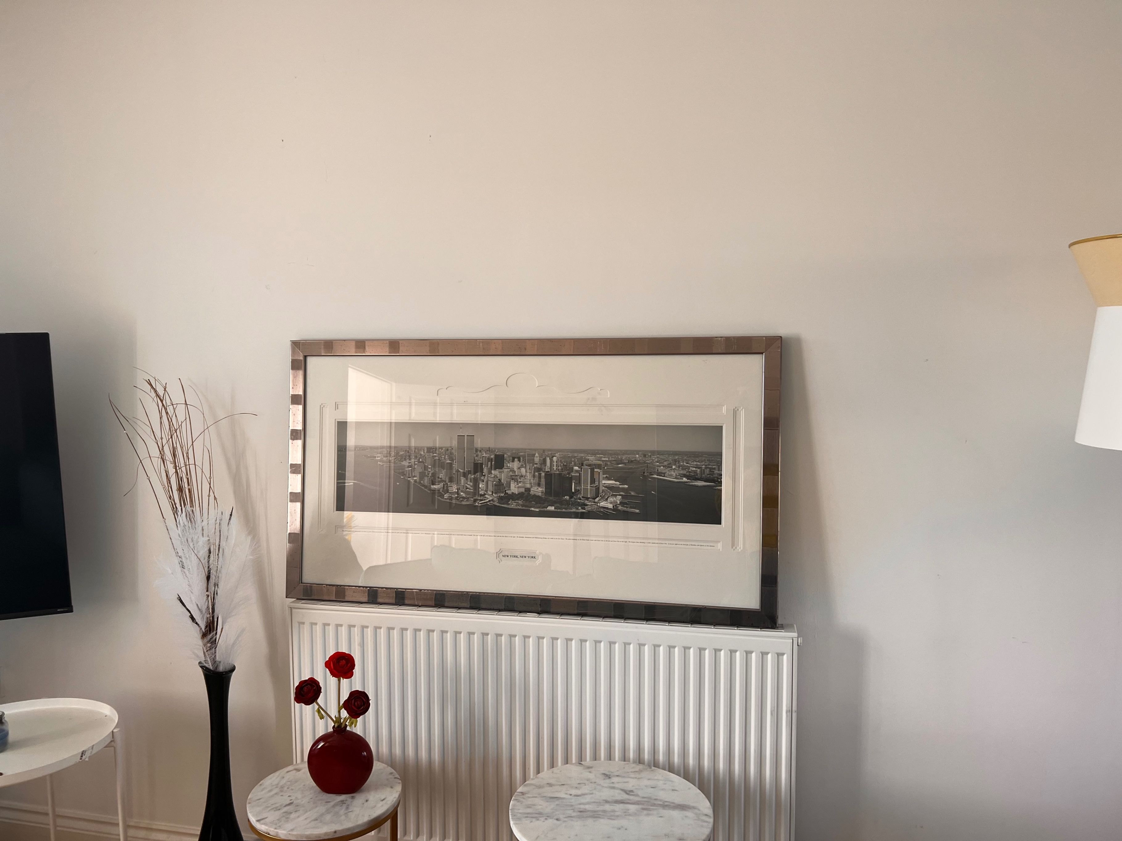 Een lijst met een foto van een stadsgezicht aan een muur, een witte radiator eronder en een vaas met rode bloemen op een bijzettafeltje.