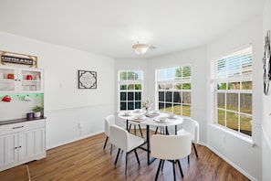 A dining area with a white table set for four, surrounded by white chairs, with a coffee bar to the left and a view of the outdoors through large windows.