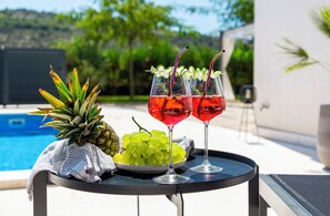 Ein Tisch mit einer Ananas, Trauben und zwei Gläsern roter Flüssigkeit, vor einem Blick auf einen Pool.