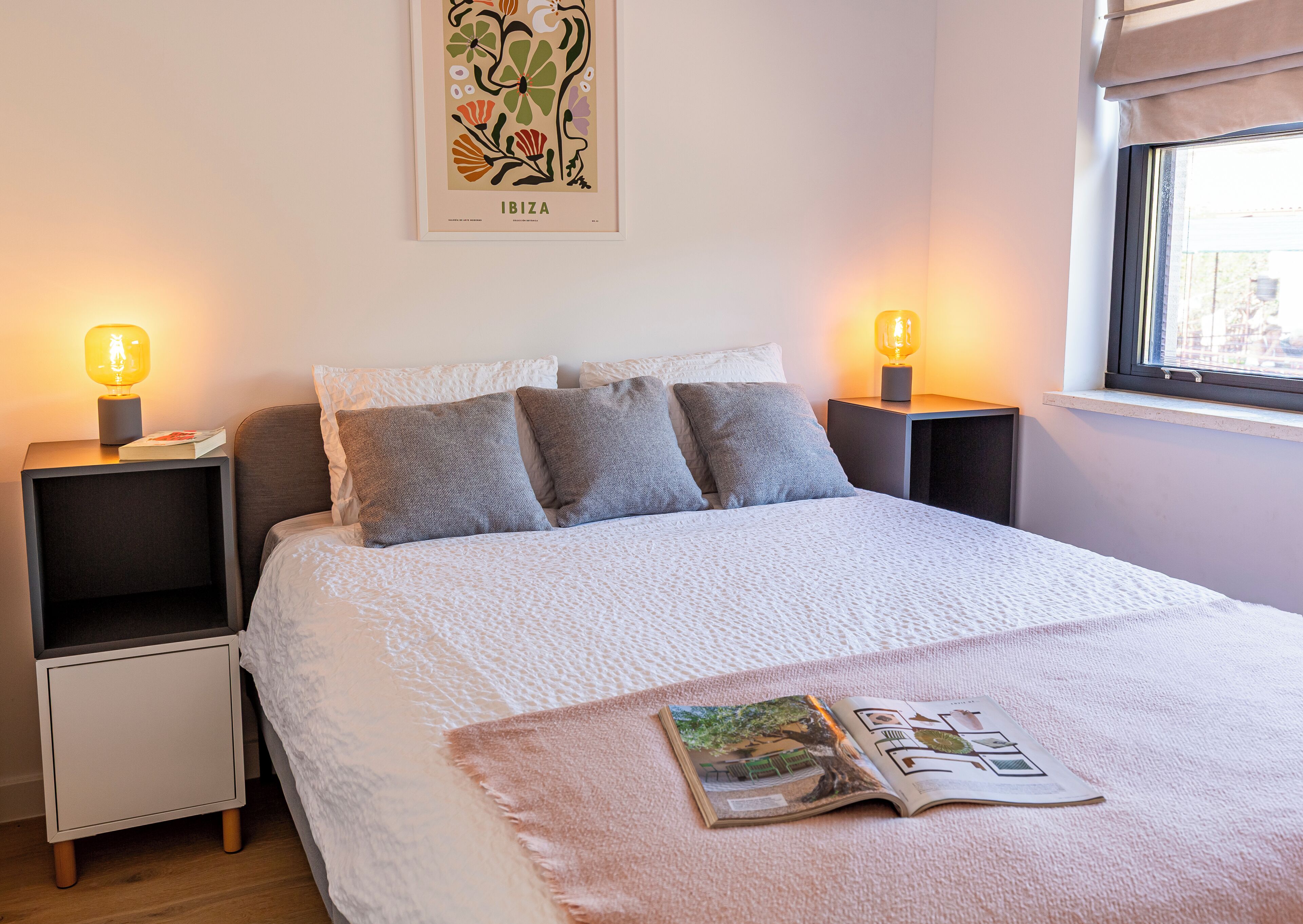 Ein ordentlich gemachtes Bett mit weißer Bettdecke, zwei Nachttischlampen und einem gerahmten Bild an der Wand.