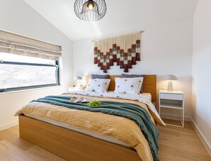 Ein Schlafzimmer mit einem hölzernen Bett, einem Nachttisch mit Lampe, einer Wandweberei und einem Fenster mit Jalousien.