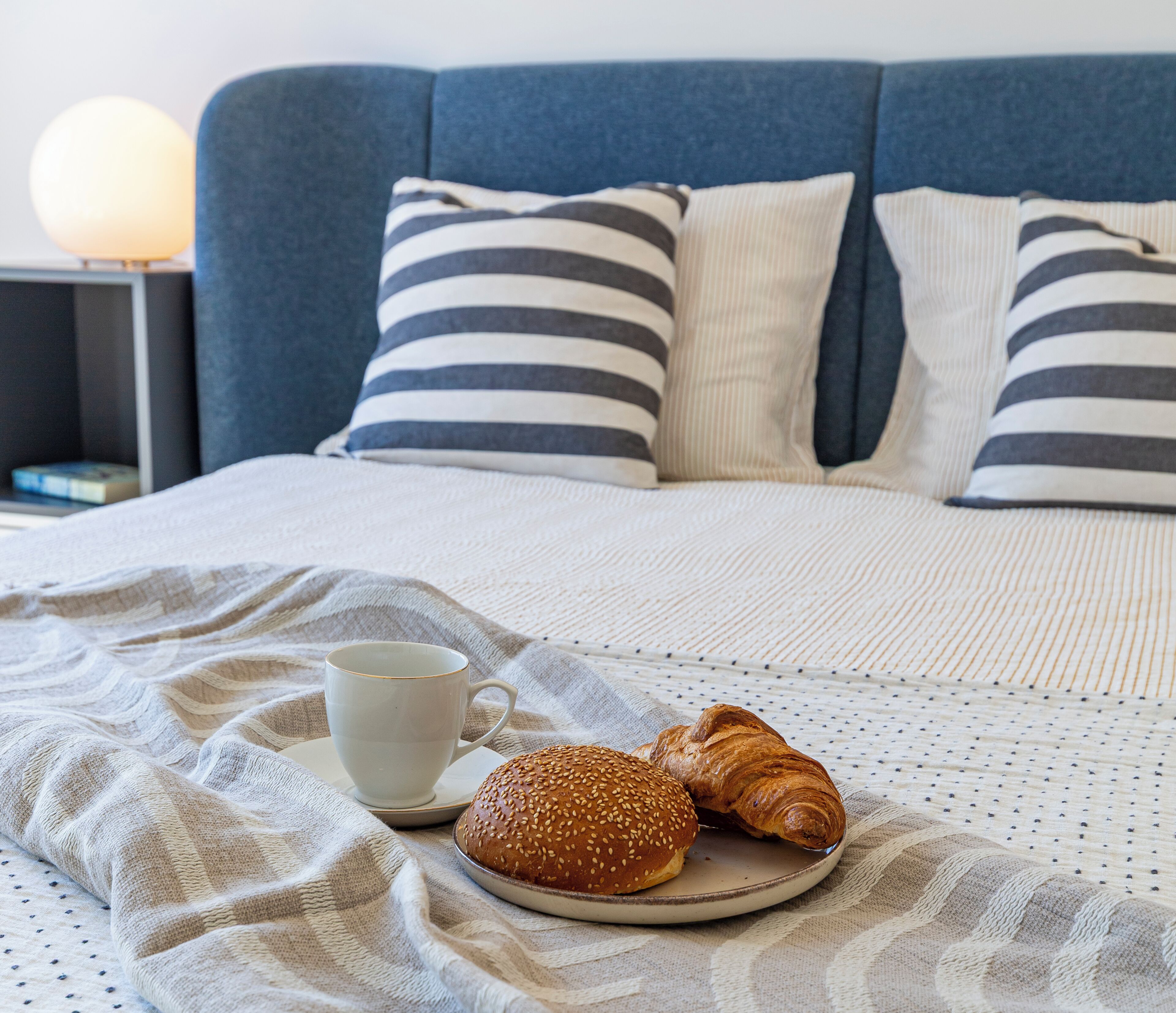 Ein ordentlich gemachtes Bett mit blauer Kopfteil, eine Tasse Kaffee und ein Croissant auf einem Teller.