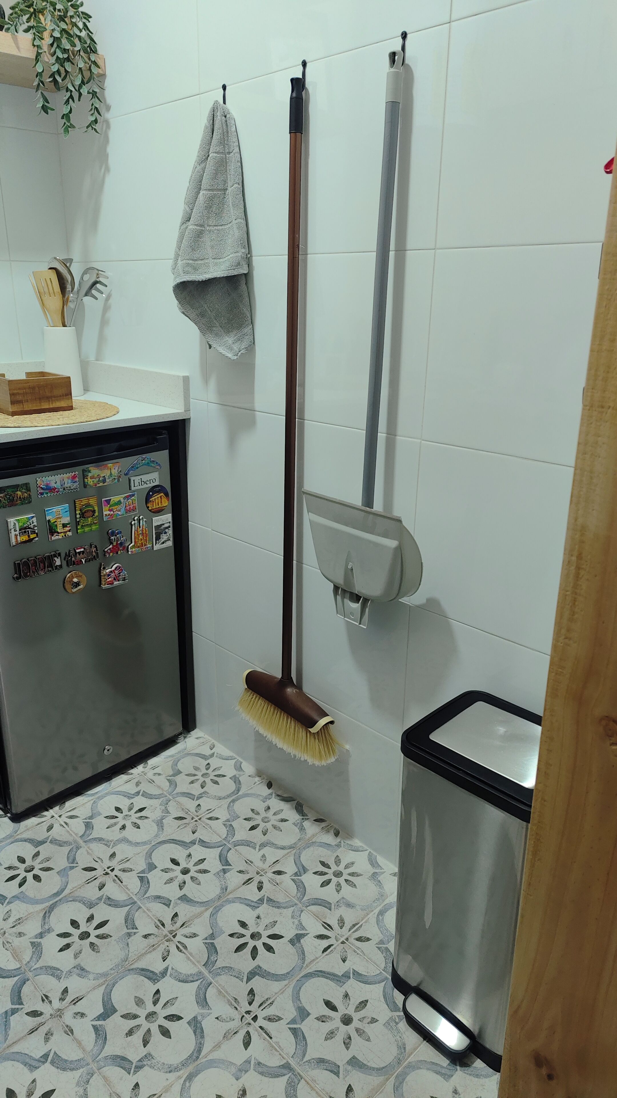A bathroom with a tiled floor, a sink with a wooden soap dish, a hanging towel, a broom, a trash bin, and a wall-mounted soap dispenser.