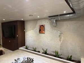 A room with a television mounted on a wooden panel, a white textured wall with a painting, and a planter with plants. - The Self-Sustainable Beach House (South Padre Island)