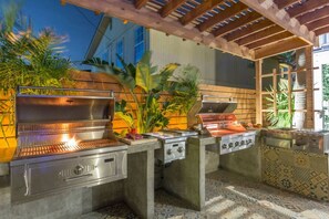 An outdoor kitchen with a grill, a sink, and a stove, surrounded by plants and a pergola. - The Self-Sustainable Beach House (South Padre Island)