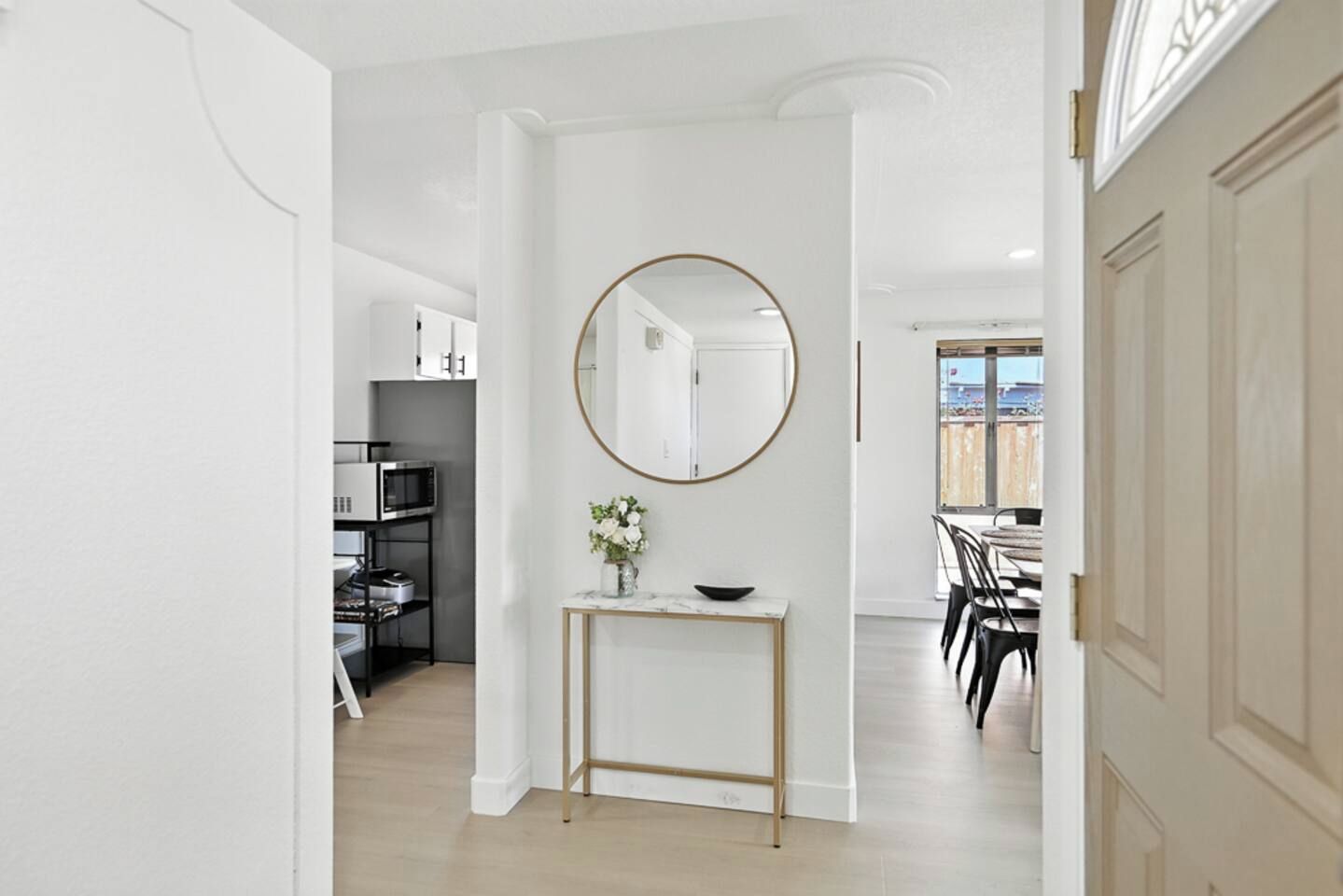 A hallway leading to a dining area with a round mirror on the wall, a small console table with a vase, and a view of the outdoors through a window.