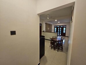 Corredor que dá para uma sala de jantar com mesa e cadeiras, ventilador de teto e janela com cortinas.