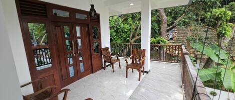 A covered outdoor seating area with wooden chairs and a white tiled floor, overlooking a lush garden.