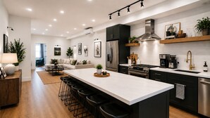 Een moderne keuken met een centraal eiland, barstoelen, roestvrijstalen apparatuur en een zithoek met een bank en stoelen.